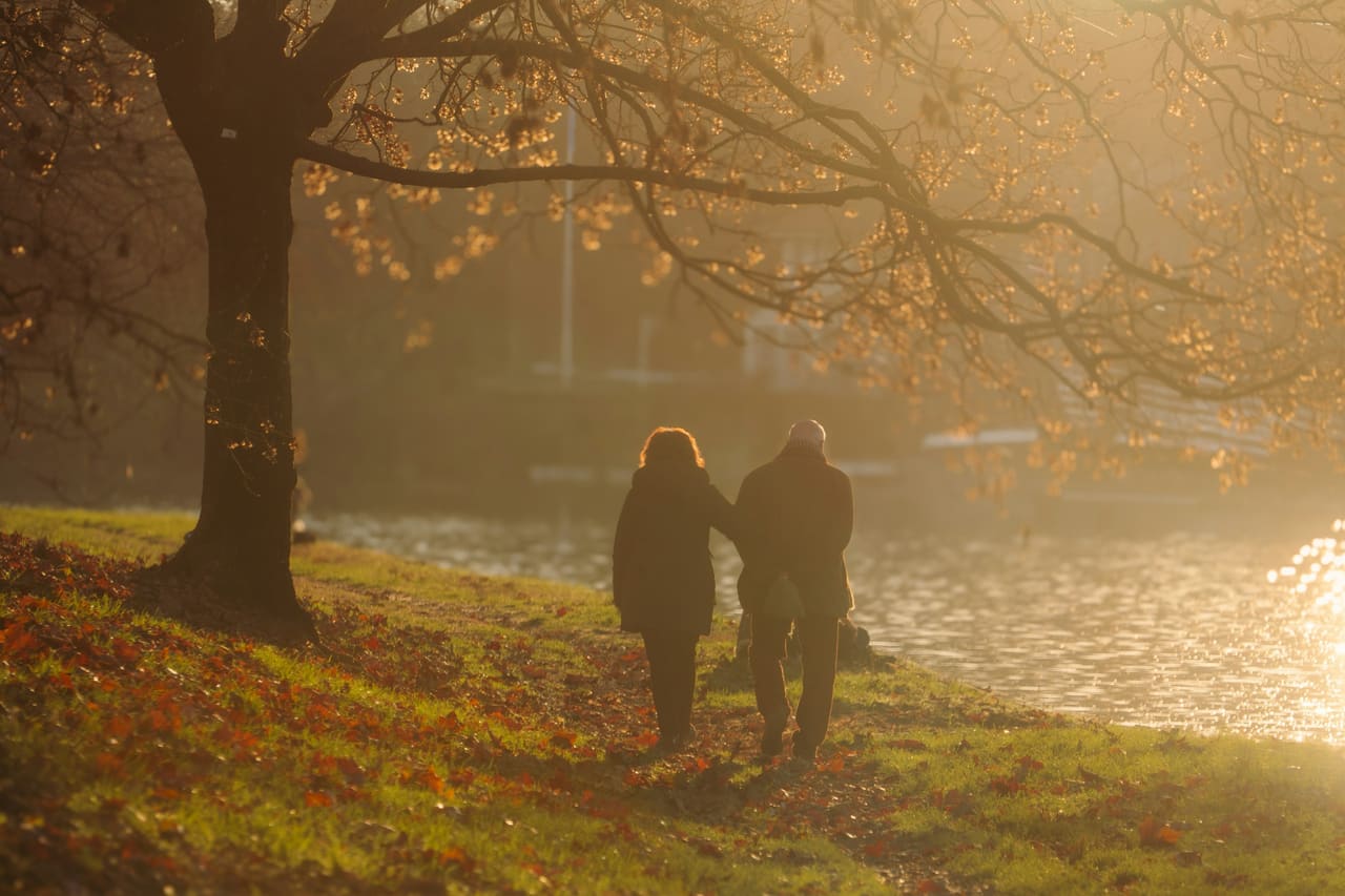 A couple walking side by side along a river path at sunset, seen from behind
