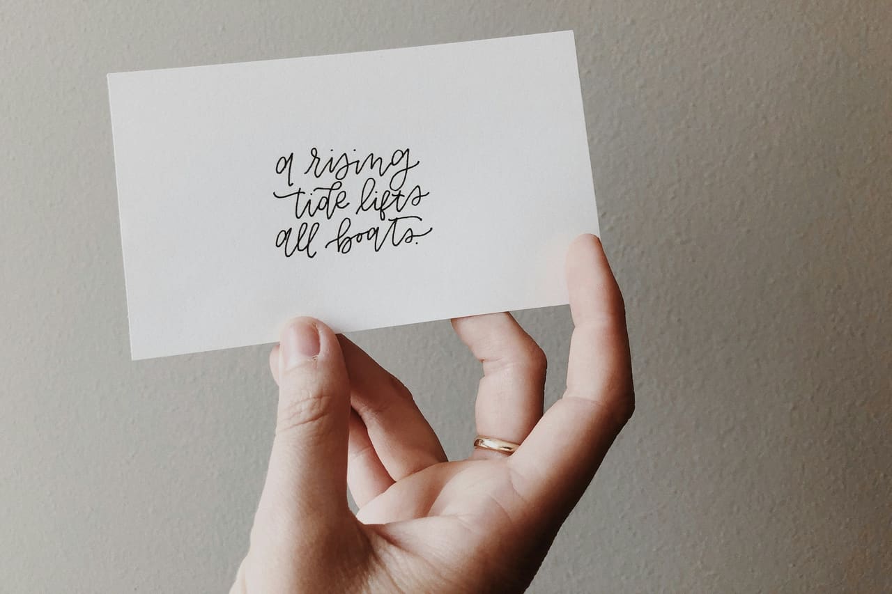 A hand holding a handwritten card that reads "a rising tide lifts all boats"
